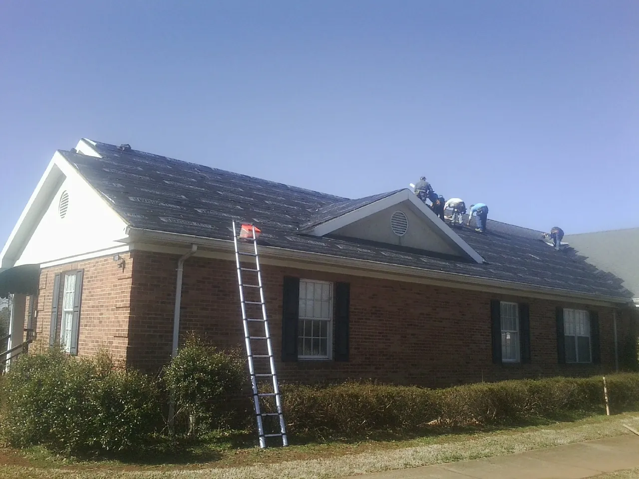 Crew tearing shingles off a brick ranch roof with a ladder set — Emergency Roof Repair work by our crew in Brookwood