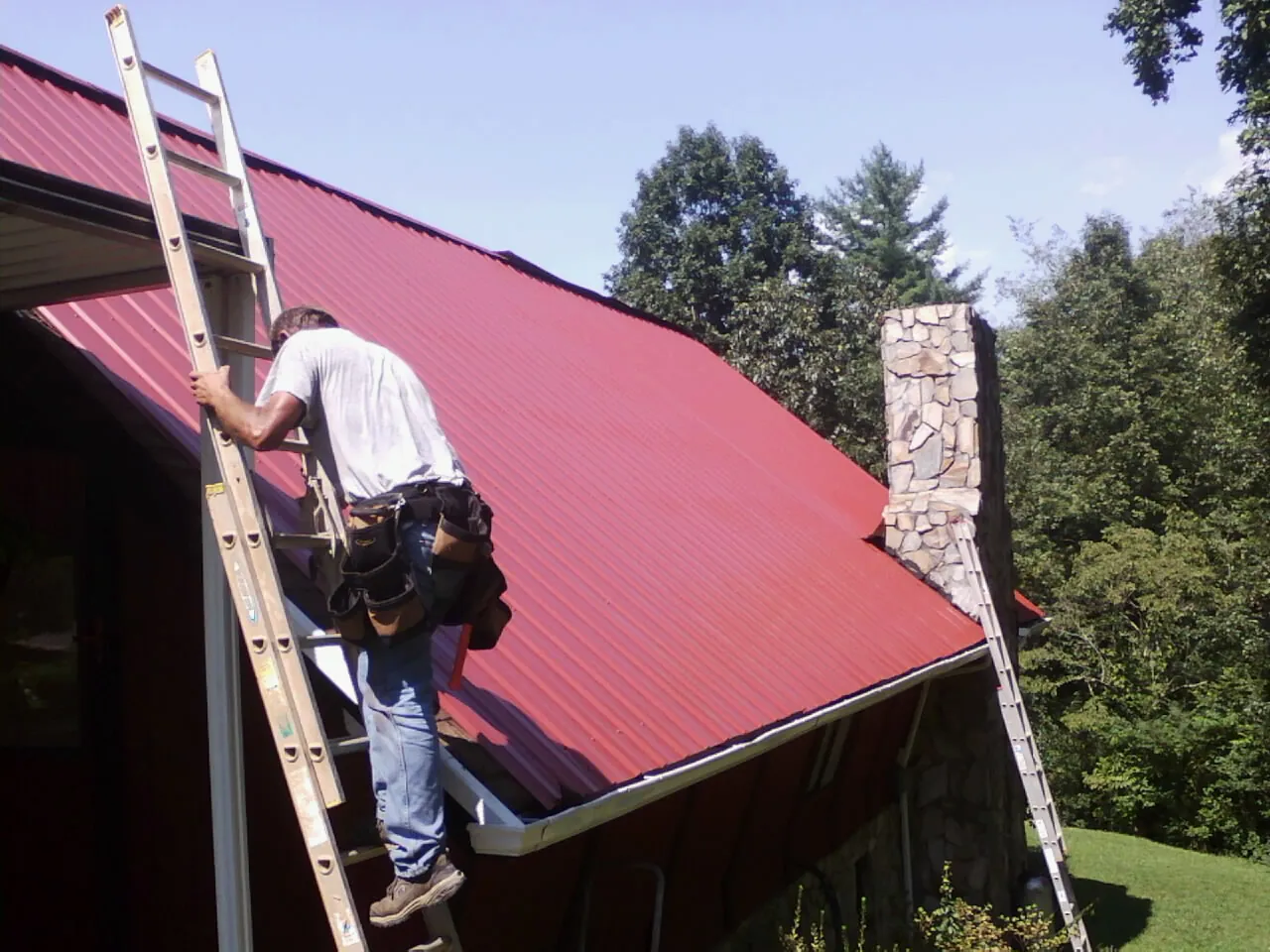 Roofer on a ladder beside a red metal roof and stone chimney — Soffit & Fascia Repair by Roofing Brothers in Brookwood